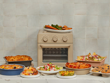 A beige toaster oven sits on a white countertop, surrounded by various baked dishes including pizza, lasagna, quiche, salads, nachos, muffins, and a platter of roasted vegetables, against a light tiled wall.