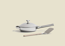 A silver frying pan with a lid and a long handle sits on a beige surface. In front of it, there is a metal spatula with a light-colored handle. The background is plain and light.