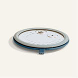 Round, flat, white and blue kitchen lid with a vent and handle, shown resting on a light-colored surface.