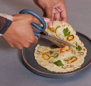 A person cutting a flatbread topped with herbs and sliced vegetables using a pair of kitchen scissors over a gray plate.