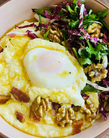A bowl of creamy polenta topped with a poached egg, walnuts, bacon pieces, and served with a side of mixed greens and herbs.