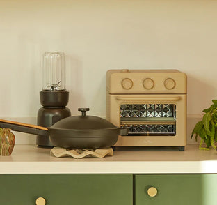 A kitchen counter with a frying pan, a blender, a beige toaster oven, a potted plant, and a small decorative object.