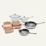 A Titanium Pro Cookware + Bakeware Set 2.0 featuring silver-toned stainless steel cookware, pale pink ceramic bakeware, and a rectangular baking sheet.