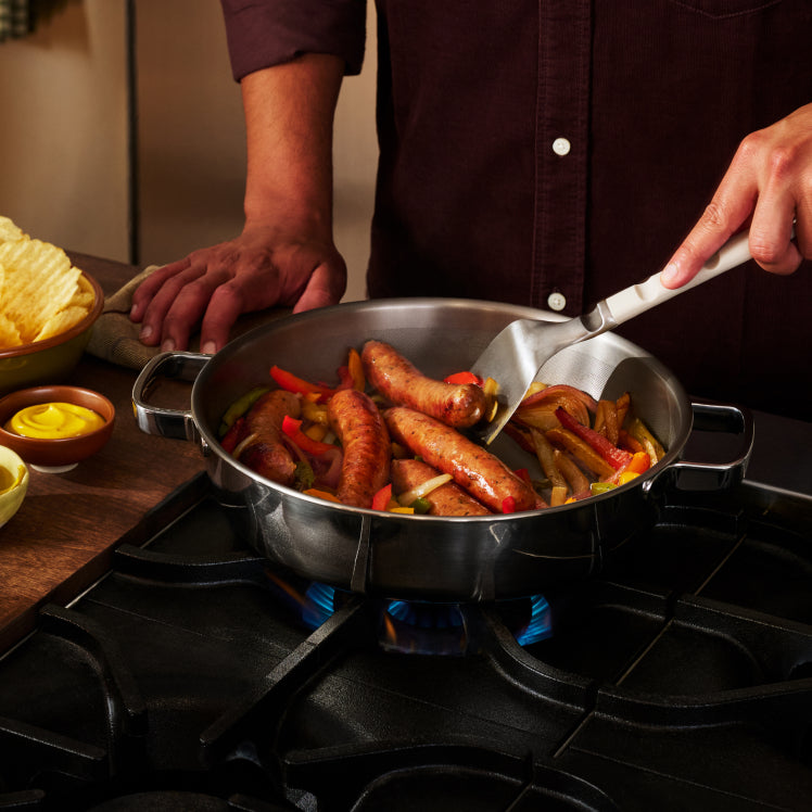 Sausages and peppers searing across the wide base of the Titanium Braiser Pro on a gas stovetop.