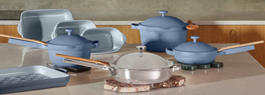 A set of modern cookware in blue and clear finishes, including pots, pans, and baking dishes with wooden handles, displayed on a kitchen counter with a light beige surface and wood cabinets in the background.