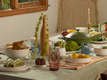 A sunlit dining table set with colorful glassware, bowls of fruit, fresh vegetables, a green Dutch oven, a lit taper candle, and decorative vases, creating a cozy, inviting meal setting.