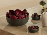 A dark grey bowl filled with red radicchio leaves sits on a beige tablecloth, next to small bowls of dates and olives, a white ceramic pitcher, and a spiky blue thistle flower.