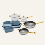 The Titanium Pro Cookware + Bakeware Set 2.0, featuring high-performance stainless steel cookware with gold-toned handles and lids, displayed alongside light and dark blue ceramic bakeware dishes.