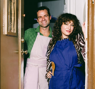 Two people standing in a doorway, one in a green shirt and apron, the other in a blue dress with a zebra-patterned cardigan.