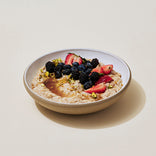 A bowl of oatmeal topped with strawberries, blueberries, blackberries, crushed nuts, and a drizzle of syrup on a light background.