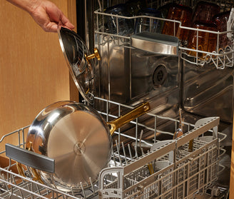 A hand places a stainless steel saucepan and its lid with gold-colored handles into the bottom rack of an open dishwasher, which is filled with various dishes and glassware.