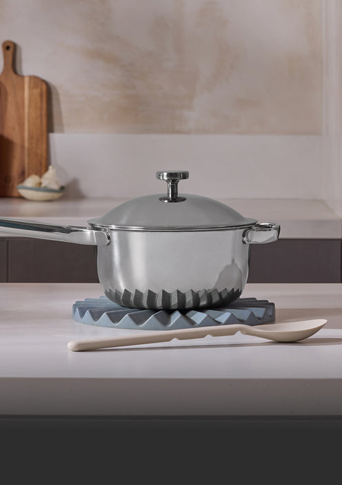 A stainless steel saucepan with a lid sits on a blue textured trivet on a kitchen counter. In front, there is a white wooden spoon. A wooden cutting board is visible in the background.