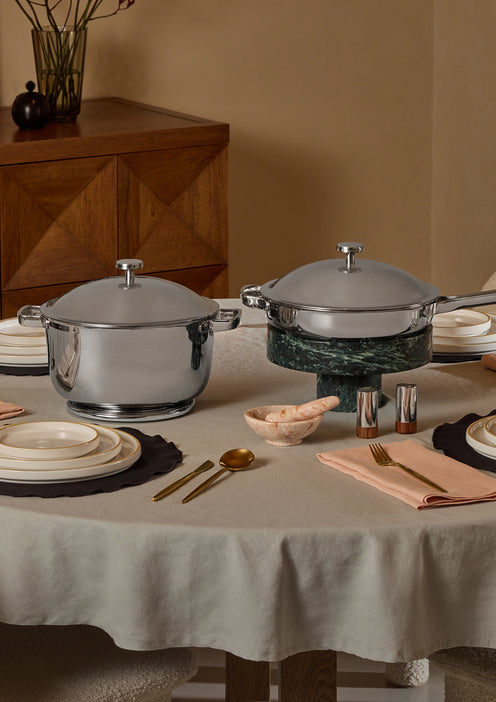 A dining table set with plates, bowls, cutlery, napkins, and two covered stainless steel pots. The table has a beige tablecloth, and a wooden cabinet with a vase and flowers is in the background.