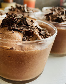 Close-up of a glass cup filled with creamy chocolate mousse, topped with dark chocolate shavings. Other similar cups are slightly blurred in the background.