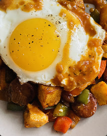 A close-up of a sunny-side up egg topped with sauce, served over a hearty mix of roasted potatoes, sausage, and colorful bell peppers.