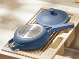 A blue nonstick frying pan with a matching lid rests on a beige dish rack over a sink. The lid has water droplets on its glass center, and the kitchen counter behind is a light, marbled surface.