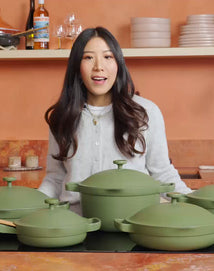 A woman with long dark hair stands in a kitchen behind a countertop displaying four green cookware pieces, including pots and pans, with shelves and kitchen items in the background.