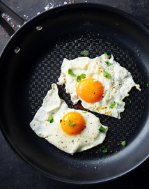 Two sunny-side-up eggs with bright yolks and sprinkled herbs cook in a black, nonstick frying pan. The eggs are seasoned with black pepper and some red flakes, with a few herbs scattered on top.