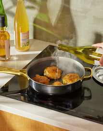 A hand pours white wine from a bottle into a skillet with seasoned chicken thighs cooking on an induction stove; wine and oil bottles are visible on the counter nearby.
