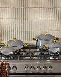 Four shiny stainless steel pots with gold-colored handles and lids sit on a modern gas stove. A brown checkered kitchen towel hangs on the oven handle, and beige tiled backsplash is visible behind.