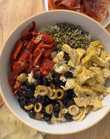 A bowl of pasta salad topped with sun-dried tomatoes, capers, artichoke hearts, black olives, and sliced green olives, set on a wooden surface next to a plate of sliced cured meats.