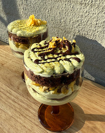 Two glass cups filled with layered desserts, featuring green cream, chocolate cake, yellow crumble, and chocolate drizzle, placed on a wooden surface in sunlight with a textured wall in the background.