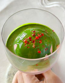 A hand holds a glass containing a smooth, round scoop of vibrant green matcha dessert topped with small red bits, possibly berries or spices, against a light background.
