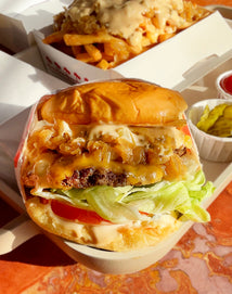 A close-up of a cheeseburger with lettuce, tomato, grilled onions, and sauce, next to a side of pickles and a tray of fries covered in melted cheese and sauce. The meal is served on a light-colored tray.