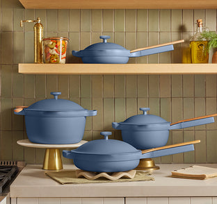 A set of matte blue cookware, including pots and pans with wooden handles and matching lids, is displayed on a kitchen counter and shelves against a tiled backsplash with jars and bottles.
