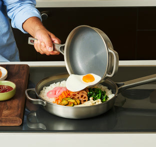 A person uses a frying pan to slide a fried egg onto a bowl of rice topped with vegetables.