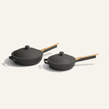 Two black nonstick pans with wooden handles and matching lids are placed side by side on a light, neutral background. One pan is slightly larger than the other.