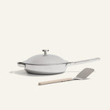 The Titanium Always Pan Pro, a white lidded frying pan with a long handle and innovative nonstick technology, sits next to a metal spatula with a beige handle on a light background.