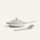 The Titanium Always Pan Pro, a white lidded frying pan with a long handle and innovative nonstick technology, sits next to a metal spatula with a beige handle on a light background.