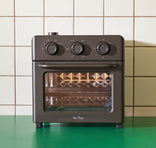 A compact, black countertop oven with three dials sits on a green surface in front of a tiled wall. The oven has a clear front door, diamond-patterned interior, and Our Place branding on the bottom.