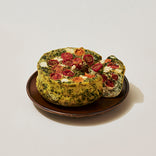 A round savory frittata with visible herbs, topped with cherry tomato slices and cheese, sits on a brown plate with a wedge cut out.
