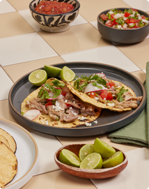 Two carnitas tacos topped with diced tomatoes, onions, cilantro, and cheese sit on a dark plate with lime wedges. Bowls of salsa, pico de gallo, and extra lime slices are nearby on a tiled surface.