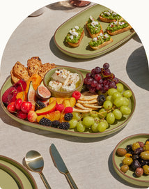 A green platter on a beige tablecloth holds assorted fruit, crackers, cheese, and a bowl of dip. Nearby are toasts with toppings, a plate of olives, and gold-colored cutlery.