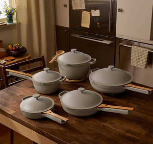 A set of grey non-stick cookware with wooden handles, including a Dutch oven, two frying pans, and a saute pan, arranged on a wooden kitchen table.