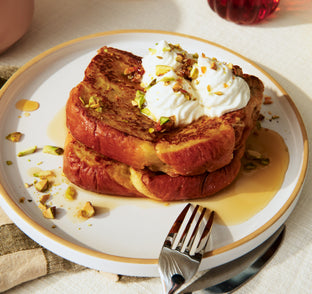 Stack of French toast with whipped cream and nuts on a white plate with gold rim, served with a side of syrup.