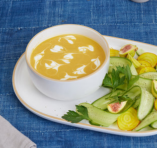 Bowl of yellow soup with a side of sliced vegetables on a blue textured tablecloth