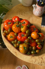 A wooden table holds a heart-shaped tray filled with ripe red and orange tomatoes and small green tomatoes. Nearby are a patterned mug with pens and part of a leafy plant. The floor and someones toes are visible at the bottom.